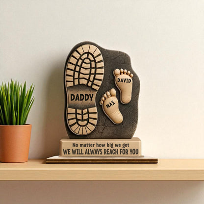 Decorative plaque with footprints and engraved text on a wooden surface with a plant in the background. Close up of personalised children names engraved on dad footprint plaque
